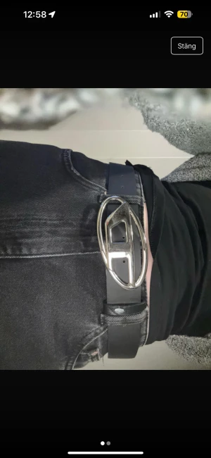 Svart bälte med ovalt silver-spänne - Snyggt svart bälte med ett stort, ovalt spänne i silverfärgad metall. Spännet har en unik design med två rektangulära detaljer och en blank finish som ger en cool vibe. Bältet är stilrent och passar perfekt till jeans eller byxor för en modern look.