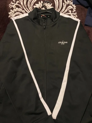 Svart Jordan vindjacka med vita detaljer - Snygg svart Jordan Air vindjacka med vita ränder längs axlar och ärmar. Jackan har dragkedja framtill, ståkrage och sportig design. Perfekt för dig som gillar streetwear och vill ha en clean look med ikonisk logga på bröstet.