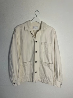 Biege overshirt från These Glory Days - Snygg biege overshirt från These Glory Days i bomull. Jackan har klassisk krage, tre stora fickor framtill och svarta knappar längs hela framsidan. Perfekt för lager-på-lager och enkel att matcha med jeans eller chinos.