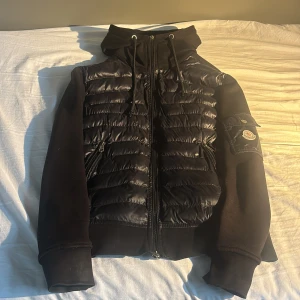 Svart pufferjacka från Moncler - Svart pufferjacka från Moncler med quiltad front i blankt material och mjuka ärmar. Jackan har huva med snörning, dragkedja framtill och Moncler-logga på ärmen. Snygg, sportig look med praktiska fickor och ribbade muddar. Storlek xs, är rätt så solblekt och det finns ett hål på armen