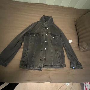 Svart jeansjacka med knappar - Säljer en svart jeansjacka med klassisk design och vita knappar framtill. Jackan har två bröstfickor med lock och två sidofickor. Snygg krage och raka, långärmade ärmar. Perfekt för dig som gillar en enkel och cool stil.