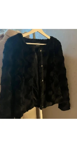 Svart pälsjacka från Vero Moda - Säljer en svart pälsjacka från Vero Moda i storlek M. Jackan har fluffig yta, rund hals och knäppning med knappar framtill. Perfekt för dig som vill ha en snygg och varm look med lite lyxig känsla. Passar dig som gillar stilrena och trendiga plagg.