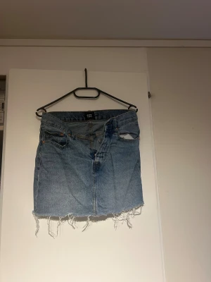 Blå jeanskjol med fransig kant från 157 - Säljer en blå jeanskjol från 157 med fransig och rå kant nedtill. Kjolen har klassiska fickor fram och bak, knapp och dragkedja i midjan. Perfekt för dig som gillar en chill och avslappnad stil. Passformen är normal och materialet är denim.