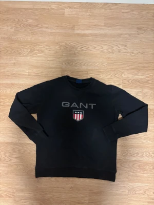 Svart sweatshirt från GANT - Svart sweatshirt från GANT med klassisk rund hals och tryckt logga samt emblem på bröstet. Tröjan har långa ärmar och ribbade muddar vid ärmslut och nederkant. Materialet är mjuk bomull som ger en skön känsla och snygg passform.