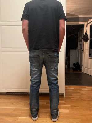 Nudie jeans  - Säljer ett par blå jeans med snygga slitningar och hål på båda knäna. Jeansen har klassisk femficksdesign, raka ben och normal passform. Färgen är mörkblå med ljusare tvättade partier och materialet är klassisk denim.