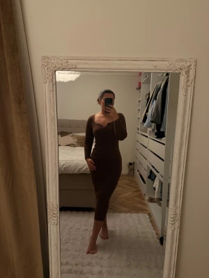 Brun ribbad långklänning - Säljer en brun, ribbad långklänning med lång ärm och v-ringad hals med vågig kant. Klänningen är figurnära och har en snygg, enkel design som känns både trendig och bekväm. Perfekt för dig som gillar stilrena plagg med det lilla extra.