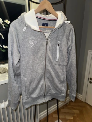 Grå hoodie från GANT med dragkedja - Snygg grå hoodie från GANT med vit huva och coolt tennisbroderi på bröstet. Tröjan har hel dragkedja, snörning i huvan och en praktisk ficka med dragkedja på sidan. Insidan är mjuk och skön, perfekt för chill eller sport.