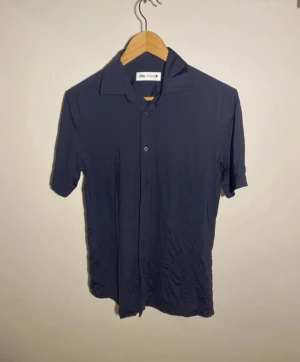 Mörkblå kortärmad skjorta från Zara - Snygg mörkblå skjorta från Zara med klassisk krage och korta ärmar. Skjortan har knappar framtill och är tillverkad i mjuk bomull som känns skön mot huden. Perfekt för dig som gillar stilrena och enkla plagg med modern passform.