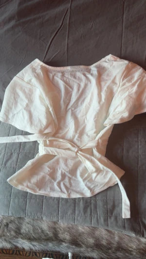 Vit omlottopp med knyt från Gina Tricot - Säljer en vit omlottopp från Gina Tricot med v-ringning och korta, lite puffiga ärmar. Toppen har knyt i midjan eller om man vill knyta den bak i ryggen. 