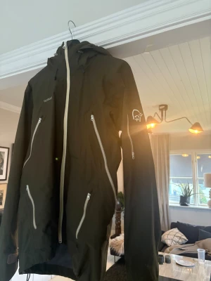Mörkgrön jacka från Norrøna - Snygg grön jacka från Norrøna med huva och flera vita dragkedjor på fram- och ärmsidan. Jackan har lång ärm, justerbar huva och coolt logotryck på armen. Perfekt för dig som gillar outdoor och vill ha en stilren jacka med funktionella detaljer.
