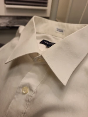 Vit skjorta med fina detaljer från Hagenfeldt L - Klassisk vit skjorta från Hagenfeldt i storlek L, tillverkad i 55% bomull och 45% polyester. Skjortan har diskret struktur med små prickar i tyget, bröstficka, långa ärmar och knäppning framtill. Perfekt för en clean och stilren look.