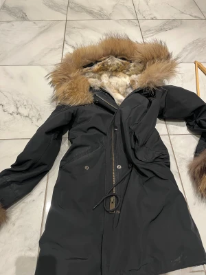 Svart parkas med pälsdetaljer - Snygg svart parkas med fluffig päls på huva, ärmslut och insida. Jackan har dragkedja framtill, snörning i midjan och flera fickor. Pälsen är i olika bruna och beige toner, vilket ger en lyxig vibe. Perfekt för kalla dagar när du vill vara både varm och stilren.
