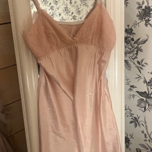 Ljusrosa satin klänning med spets - Supersöt ljusrosa klänning i satin med spets upptill och smala axelband. Klänningen har en v-ringad urringning och ärmlös design, vilket ger en feminin och mjuk look. Perfekt för dig som gillar romantiska detaljer och skön passform.