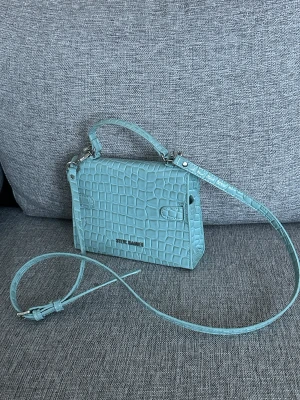 Turkos axelväska från Steve Madden - Snygg turkos axelväska från Steve Madden med croco-mönstrad yta och glansig finish. Väskan har både handtag och justerbar axelrem, samt silverfärgade metalldetaljer och lås framtill. Perfekt storlek för det viktigaste och ger en cool touch till din outfit.