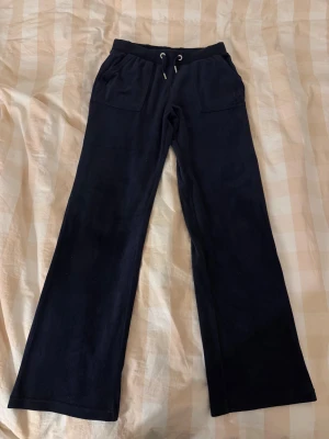 Mörkblå mjukisbyxor Juicy Couture - Mörkblå mjukisbyxor från Juicy Couture med raka ben och snörning i midjan med silverfärgade detaljer. Byxorna har stora fickor fram och bak, samt broderad JC-logga på ena fickan. Mjukt och skönt material som ger en chill vibe.
