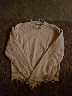 Beige stickad tröja från Bershka - Säljer en beige stickad tröja från Bershka i storlek S. Tröjan har långärmade ärmar och råa, slitna detaljer vid halsringning, muddar och nederkant för en edgy look. Perfekt för dig som gillar streetstyle och vill ha något unikt i garderoben.