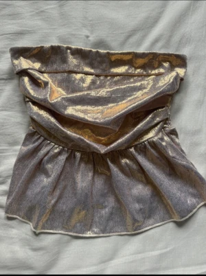 Glittrig guld bandeau tubtopp - Säljer en glittrig bandeau tubtopp i guld med volangkant nedtill. Toppen har en snygg rynkad framsida och smockad baksida för extra komfort. Materialet är mjukt och skimrande, perfekt för att sticka ut. Passar dig som vill ha en unik och trendig topp med metallic vibe. (Aldrig använd, står ej storlek men passar mig som har small)