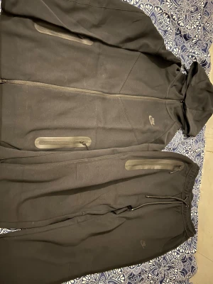 Svart Nike träningsset med huva M - Nike svart träningsset i storlek M, består av hoodie med dragkedja och huva samt matchande byxor med dragsko. Båda delarna har diskreta Nike-loggor och praktiska fickor med dragkedja. Perfekt för chill eller träning, mjukt material och sportig look. Fick den nyligen i present men den var för stor. Har bara använt den 2 gånger 