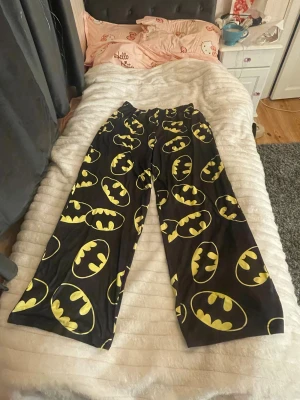 Batman-mönstrade svarta mjukisbyxor och T-shirt  - Svarta mjukisbyxor med stort Batman-tryck i gult över hela byxorna. Byxorna har en loose passform och elastisk midja för extra komfort, säljer även med T-shirten till som har ett Batman tyck på bröstet som också är i storlek L som byxorna. 