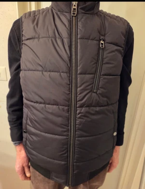 Svart quiltad dunväst från Jofama - Svart dunväst från Jofama med quiltad design och hög krage. Västen har en snygg bröstficka med dragkedja och en kraftig dragkedja framtill. Perfekt att slänga över en hoodie för extra värme. Stilren look och passar dig som gillar klassiska plagg. Nytt pris: 2000kr