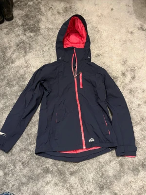 Mörkblå vindjacka från McKinley - Snygg mörkblå vindjacka från McKinley med rosa detaljer och dragkedjor. Jackan har huva, lång ärm och reflexdetalj på ryggen. Perfekt för blåsiga dagar och outdoor-aktiviteter. Materialet är lätt och syntetiskt, med smarta fickor och justerbara muddar.