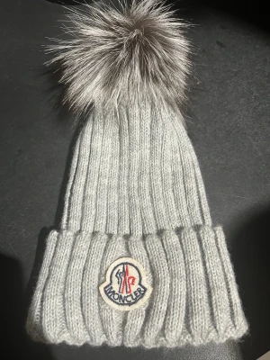 Grå Moncler mössa med pälsboll - Snygg grå ribbstickad mössa från Moncler med stor fluffig pälsboll på toppen. Märkets logga är broderad framtill på uppviket. Perfekt för dig som vill ha en lyxig och varm accessoar med streetkänsla.