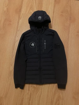 Svart Sail Racing dunjacka med huva - Helt ny sailracing jacka, fick den som present nypris på den var runt 4299kr, men den kom inte till användning. Har testat på mig den 2 gånger. Skicket på den är precis som nyköpt, bara att den saknas tags.