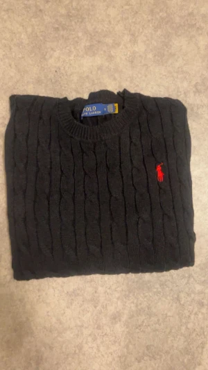 Svart kabelstickad tröja Polo Ralph Lauren - Svart kabelstickad tröja från Polo Ralph Lauren i storlek S. Klassisk rund hals och långärmad modell med röd broderad logga på bröstet. Tröjan är stickad i mjukt material och har ribbade muddar vid ärmslut och nederkant.inga skador.