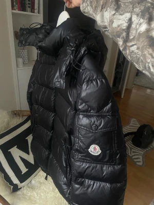 Svart Moncler pufferjacka med huva - Säljer denna jacka som jag inte vet är äkta, därav priset. jackan har inga defekter och dragkedjan funkar bra. Det står ingen storlek men skulle säga att den är S eller M. Jag bär den lite overs size och är en storlek. S. Den är i äkta dun och mycket varm. 