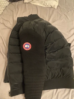 Svart pufferjacka från Canada Goose - Säljer en svart pufferjacka från Canada Goose med stickade ärmar och klassisk logga på vänster arm. Jackan har två dragkedjeförsedda fickor fram, ribbade muddar och hög krage. Perfekt för kalla dagar och har en modern, clean look. Inte använt alls mycket passar både s och m 