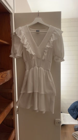Vit volangklänning med v-ringning - Supersöt vit kortklänning med v-ringning och volangdetaljer längs axlar och ärmar. Puffärm med resår och smock i midjan. Använd 2 ggr storlek XS från Gina❤️
