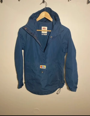 Blå Fjällräven Vardag Anorak XS - Säljer en blå Fjällräven Vardag Anorak med huva och dragkedja i sidan. Jackan har en klassisk anorak-look med stor ficka framtill och justerbar dragsko nedtill. Perfekt för dig som gillar outdoor och streetstyle.