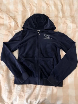Juicy Couture mörkblå velour hoodie - Mörkblå hoodie från Juicy Couture i mjuk velour med glittrande strasslogga på bröstet och stor Juicy Couture-text på ryggen. Jackan har huva, två fickor fram och dragkedja med J-formad zip. Perfekt för dig som gillar ikonisk Y2K-stil eller bara chill vardagsstil.