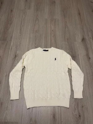 Vit kabelstickad tröja Polo Ralph Lauren - Vit kabelstickad tröja från Polo Ralph Lauren med klassisk rund hals och svart broderad logga på bröstet. Tröjan har långärmad passform och ribbade muddar vid ärmslut och nederkant. Perfekt för dig som gillar stilrena och tidlösa plagg.