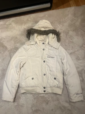 Vit pufferjacka College Athlete - Snygg vit pufferjacka från College Athlete med huva och fuskpälsdetalj. Jackan har ribbade muddar, två fickor med knappar och både dragkedja och knappstängning framtill. Broderad logga på bröstet och ärmen. Perfekt för kalla dagar. Storlek 146