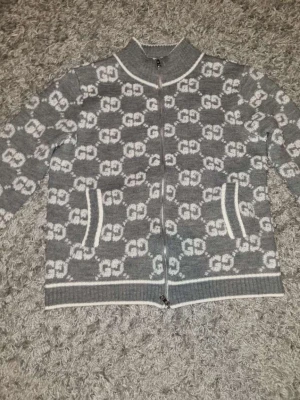Grå stickad cardigan jacka från Gucci - Säljer en grå stickad cardigan jacka från Gucci med vit GG-logga över hela jackan. Jackan har dragkedja framtill, ribbade muddar och vita detaljer vid krage, fickor och nederkant. Perfekt för dig som vill ha en snygg och trendig look. Pris går att diskutera.