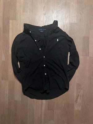 Svart skjorta från Ralph Lauren M - Svart långärmad skjorta från Ralph Lauren i featherweight mesh-bomull. Klassisk passform med knappar framtill och vit broderad logga på bröstet. Perfekt för dig som gillar stilrena och tidlösa plagg.  Passar för runt 184 🙌  9/10 skick ✅ Skickar inom 48 timmar om inte samma dag 🚚