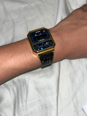 Casio Pac-Man digital klocka guld/svart - Den köptes för 6000kr. En unik digital klocka från Casio med Pac-Man tema. Rektangulärt guldigt hölje och svart metallarmband med Pac-Man-mönster och figurer. Displayen har blå och röda detaljer, och armbandet är i rostfritt stål. Perfekt för dig som gillar retro och gaming. Limited Edition.