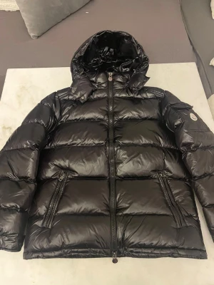 Svart Moncler pufferjacka med huva - Säljer en svart pufferjacka från Moncler med glansig finish och stor huva. Jackan har dragkedja framtill, två snedställda fickor med dragkedja och en ficka med Moncler-logga på ärmen. Perfekt för kalla dagar och riktigt snygg streetstil.