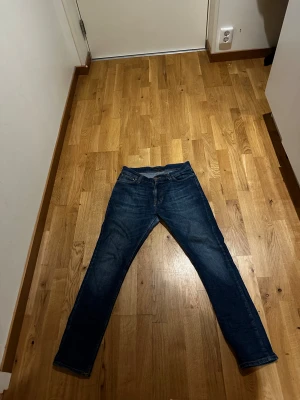 Blå Nudie Jeans Skinny 28/30 - Säljer ett par blå skinny jeans från Nudie Jeans i storlek 28/30. Klassisk femficksmodell med orange sömmar och Nudie-logga på bakfickan. Jeansen har en smal passform och är tillverkade i mjukt denimtyg som sitter snyggt på benen.