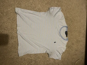 Randig t-shirt från Polo Ralph Lauren - Snygg t-shirt från Polo Ralph Lauren i ljusblått och vitt med smala ränder. Klassisk passform med korta ärmar och rund halsringning. Liten broderad logga på bröstet. Tillverkad i mjuk bomull och modellen är slim fit alltså liten i modellen.