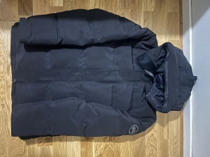 Svart pufferjacka från Canada Goose - Säljer en svart pufferjacka från Canada Goose med huva och diskret logotyp på ärmen. Jackan har en stilren look, dragkedja framtill och är tillverkad i slitstark polyester. Perfekt för kalla vinterdagar och har en normal passform. Storlek L men den passar mer M 