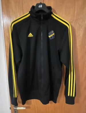 Svart AIK Adidas ziptröja M i mycket bra skick - Svart ziptröja från Adidas med AIK-logga på bröstet och klassiska gula ränder längs ärmarna. Tröjan har hög krage, lång ärm och dragkedja framtill. Perfekt för dig som gillar sportig stil och vill visa ditt AIK-support. Materialet är polyester.