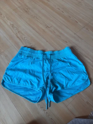 Blå och rosa sportshorts från H&M, strl 36 - Säljer ett par ljusblå och neon rosa sportshorts från H&M Sport i storlek 36. De har elastisk midja med snörning och meshfoder på insidan för extra komfort. Perfekta för träning eller aktiva dagar. Materialet är lätt och syntetiskt, vilket gör dem riktigt sköna att bära.