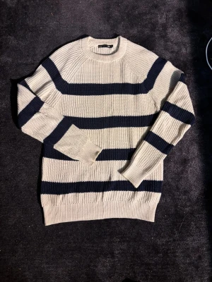 Randig stickad tröja i beige och marinblå - Säljer en grovstickad tröja med breda marinblå och beige ränder. Tröjan har rund halsringning, lång ärm och ribbade muddar. Perfekt för dig som gillar en chill och klassisk look med lite nautisk vibe.