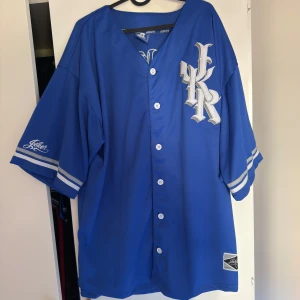 Blå Joker baseballskjorta med tryck xxl - Snygg blå baseballskjorta från Joker med stora vita tryck på bröstet och ryggen, där det står 'Joker' och siffran 69. Skjortan har vita knappar framtill, breda korta ärmar med vita ränder och coolt LA-inspirerat märke. Perfekt för dig som gillar streetwear och sportig stil.