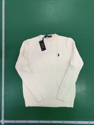 Vit kabelstickad tröja Polo Ralph Lauren - Vit kabelstickad tröja från Polo Ralph Lauren i storlek L. Tröjan har rund hals, långärmad design och klassisk broderad logga på bröstet. Mjuk och skön känsla med ribbade muddar och nederkant. Perfekt för dig som gillar stilren och tidlös look.