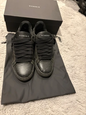 Svarta sneakers från Numeris med vita stänk - Svarta sneakers från Numeris med coola vita färgstänk på både ovandel och sula. Skorna har rund tå, platt sula och breda svarta snören. Ovandelen är i skinn med textilfoder och detaljerade sömmar. Perfekt för dig som gillar en unik och urban stil. Men har lite slitage på hälarna på båda skorna men det är inte synligt.