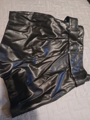 Svarta skinnshorts från H&M Divided - Säljer ett par svarta shorts i syntetiskt skinn från H&M Divided, storlek 34. De har hög midja, breda hällor och medföljande bälte med metallring. Shortsen har dragkedja i sidan och är fodrade för extra komfort. Perfekt för dig som gillar edgy och trendig stil.