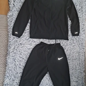 Svart Nike vindjacka och byxor(dress) med reflexdetaljer - Svart vindjacka från Nike med hel dragkedja, reflekterande detaljer och två sidofickor med meshfoder. Jackan har en sportig look och är tillverkad i lätt syntetmaterial, perfekt för träning eller när du vill ha en clean stil. Klassisk Nike Swoosh på bröstet.